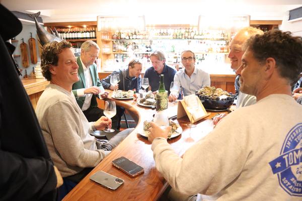 A group of men are gathered around a wooden table in a lively restaurant or bar, enjoying oysters and white wine